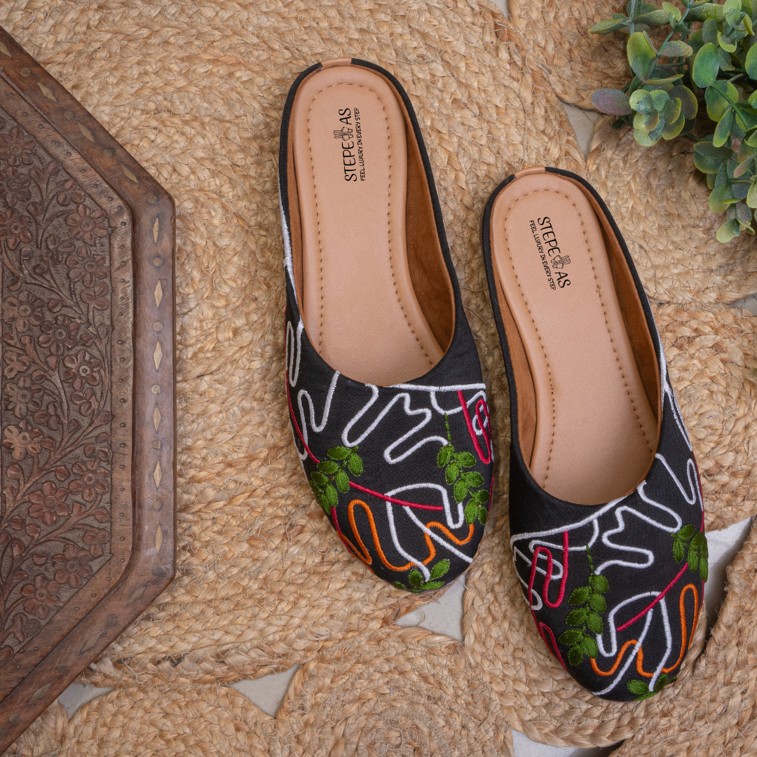 Stepellas black juttis featuring vibrant green, orange, and pink floral embroidery with white vine patterns.