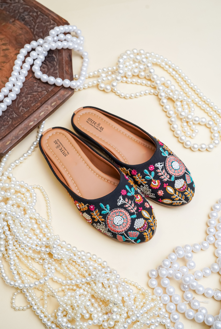 Stepellas black ethnic juttis featuring vibrant pink and yellow floral embroidery with green vine patterns.