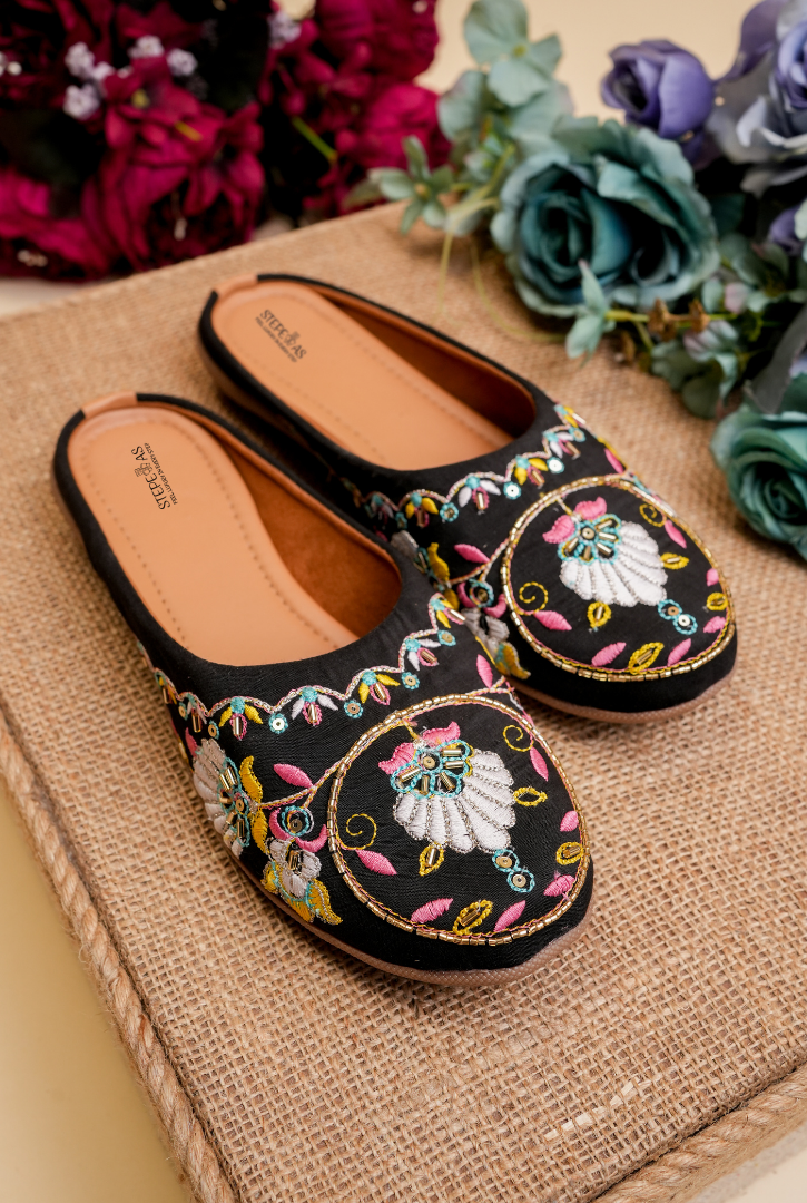 Stepellas black juttis featuring a large round mandala-style embroidery with pink and yellow threadwork.