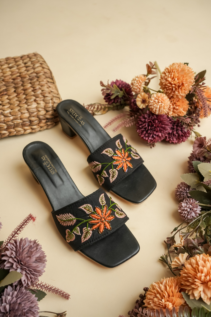 A pair of luxury Stepellas branded black heels with vibrant handcrafted orange flower embroidery on an aesthetic background.