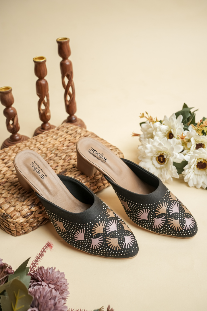 A pair of luxury Stepellas branded black heels with intricate round stone work displayed with aesthetic floral decor.